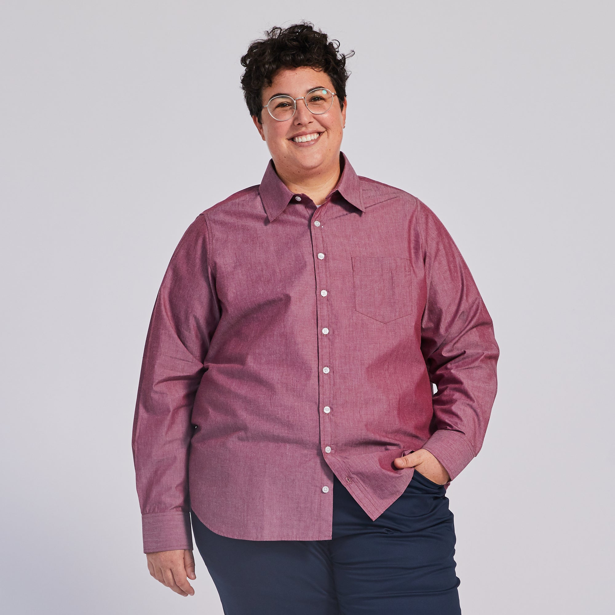 A person smiling and wearing a red chambray point collar shirt with buttons down the front. Color: Red Chambray