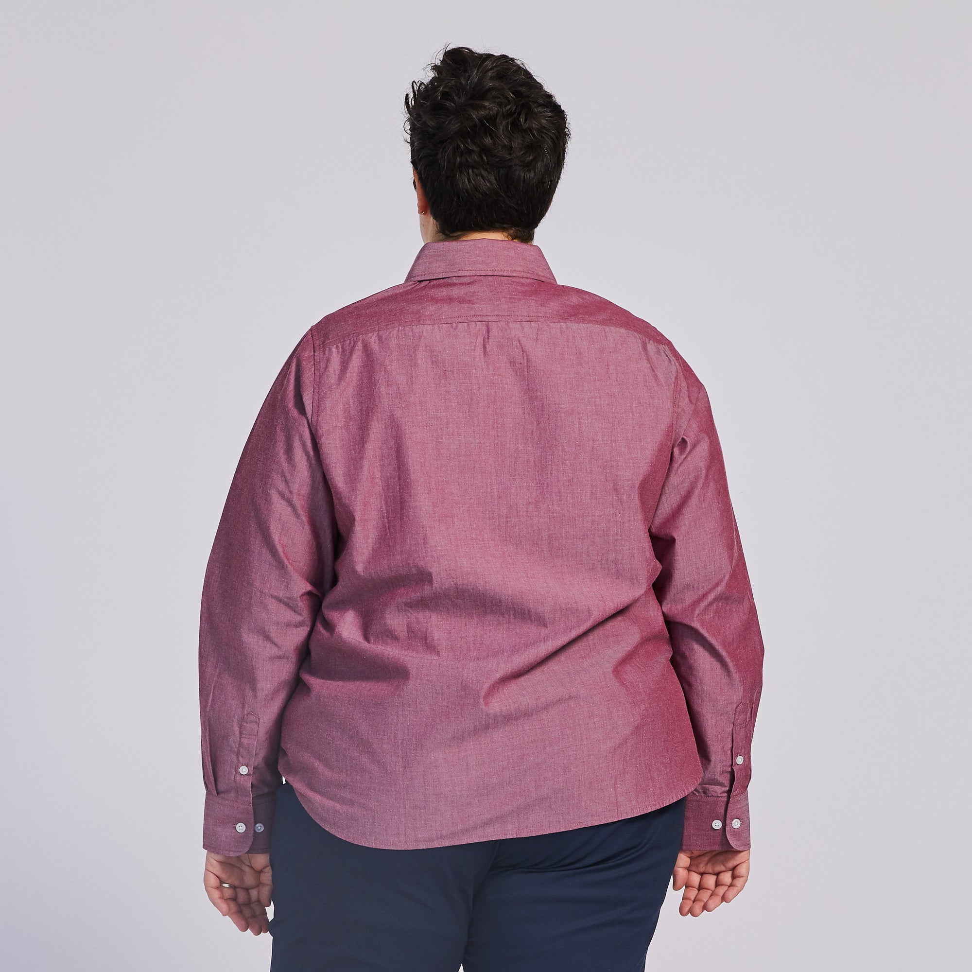 A person facing a wall, wearing a chambray point-collar shirt. Color: Red Chambray