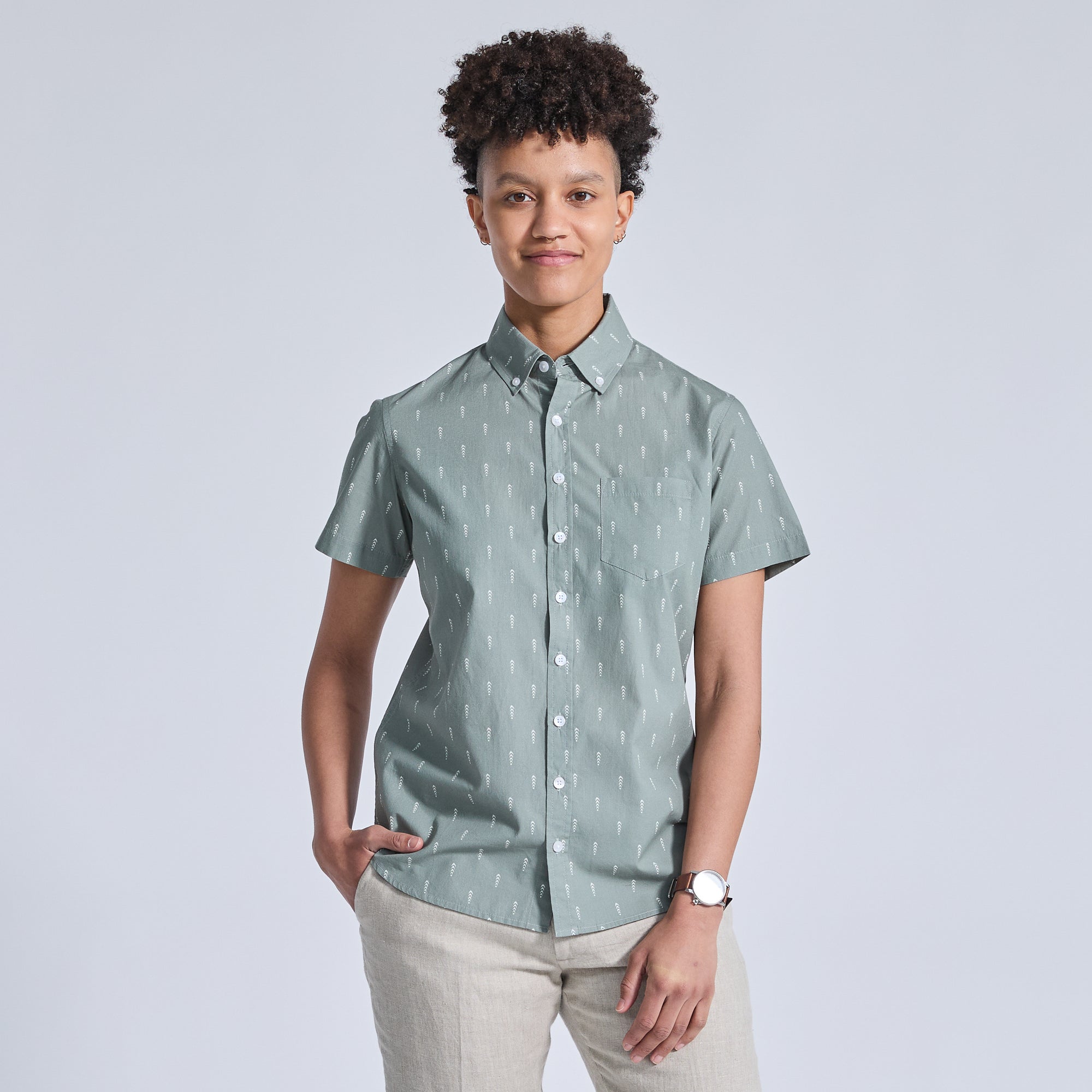 A person wearing a short-sleeved, button-down shirt in a green color with a geometric pattern, paired with light-colored pants. Color: Cypress