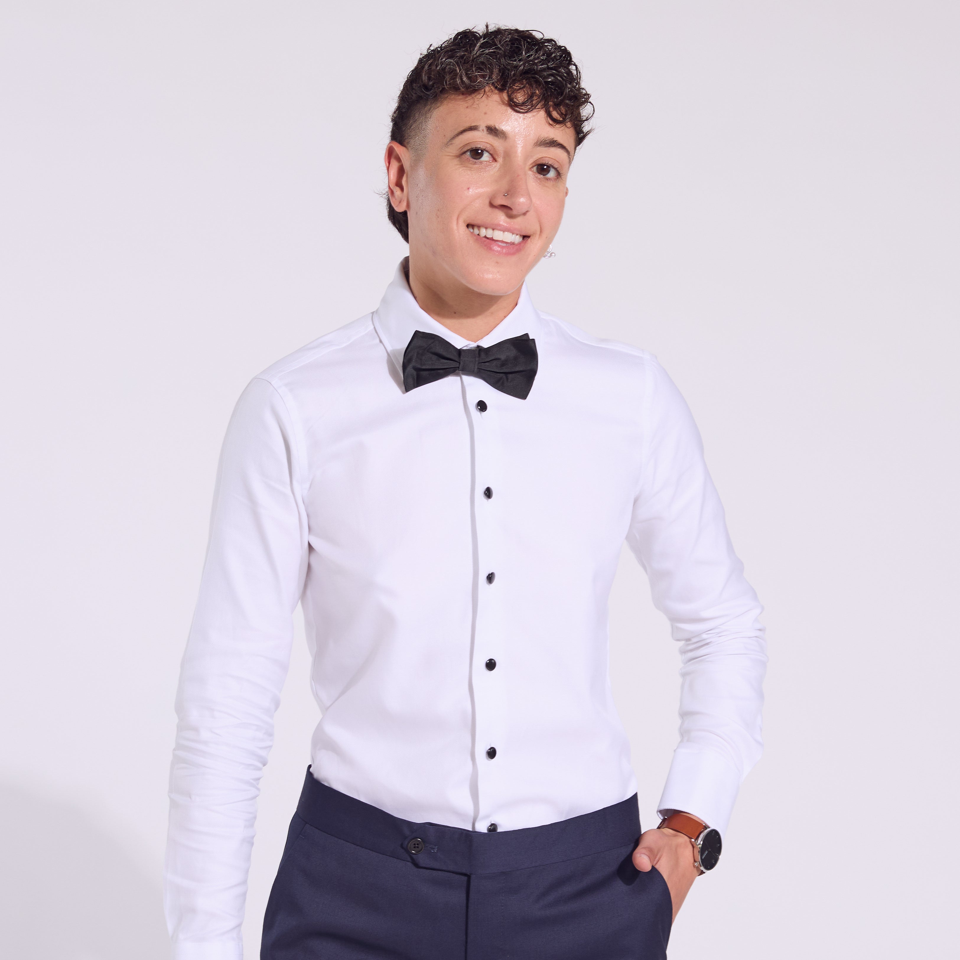 Person wearing a white dress shirt with black buttons and a black bow tie, standing against a plain background.