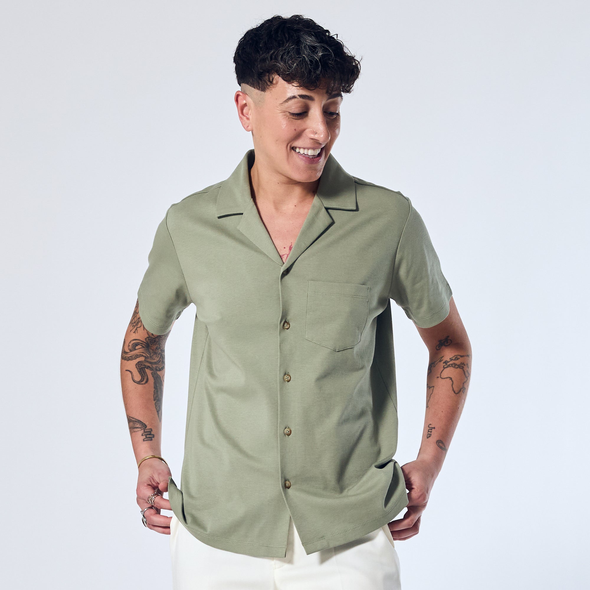 Androgynous model wearing a short-sleeved sage green camp collar shirt with a chest pocket and faux horn buttons, paired with white pants. Color: Sage
