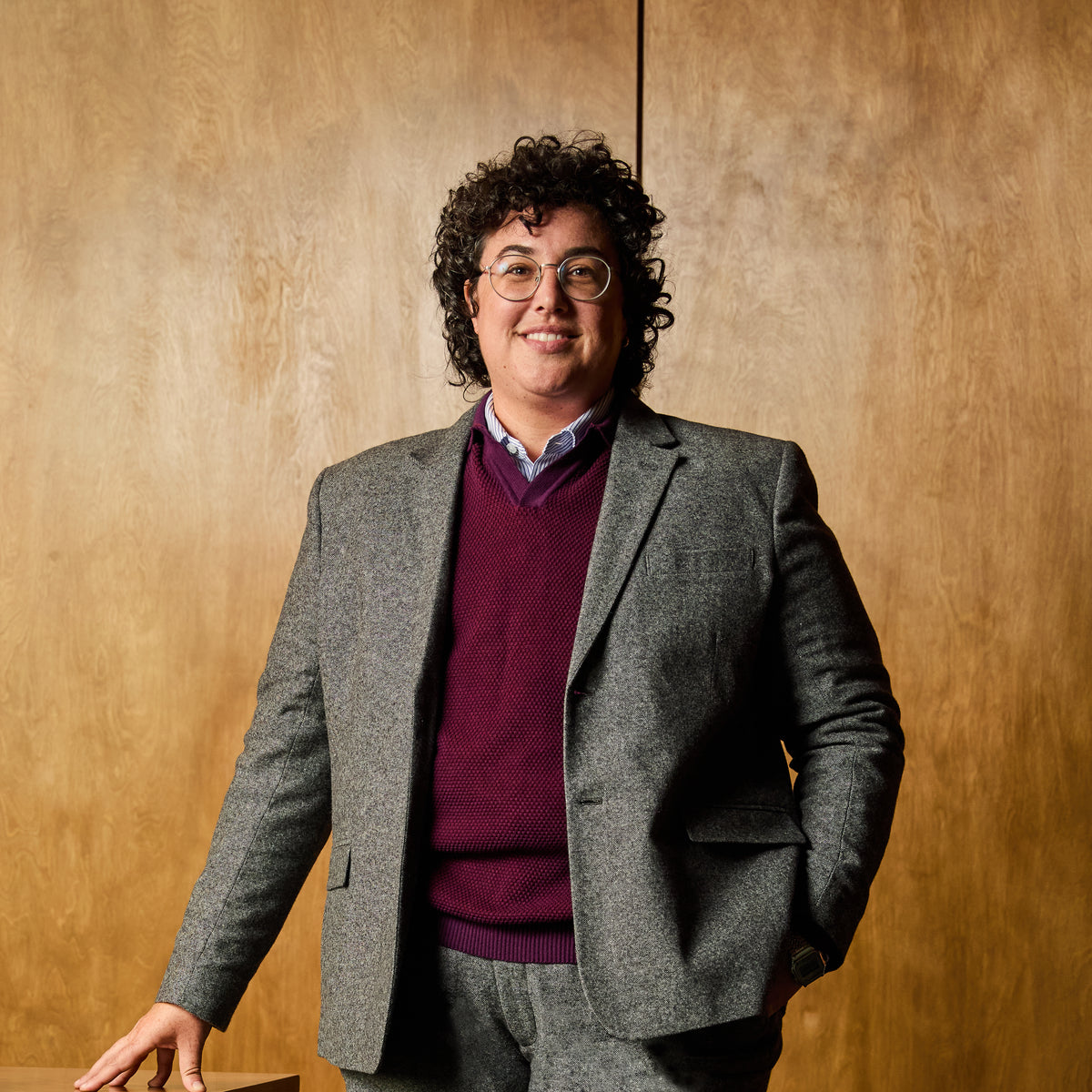 Person wearing a gray blazer and purple sweater against a wooden background.  Color: Smokey