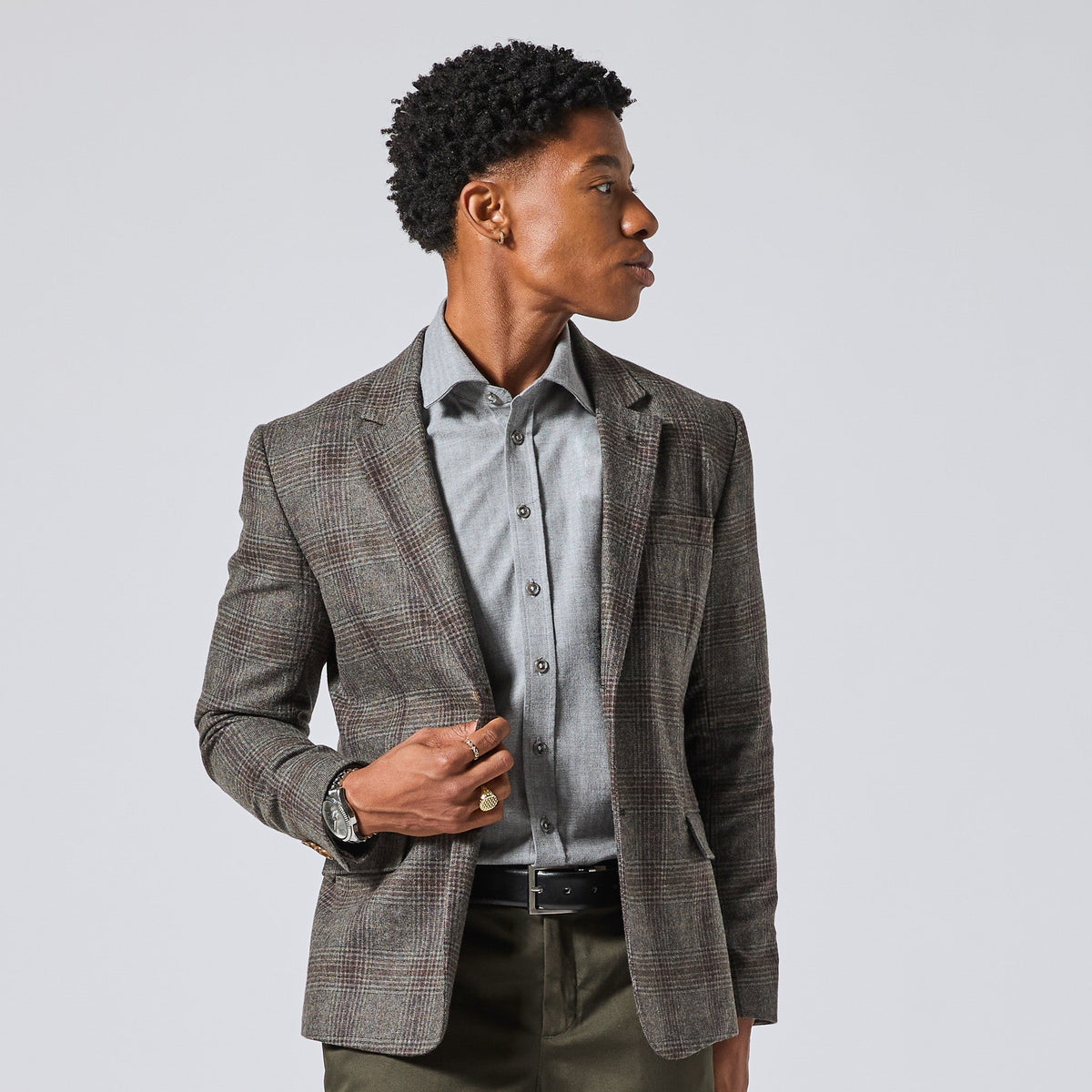 Model wearing a gray and brown plaid blazer over a light gray button-down shirt, styled with olive green trousers, against a solid light gray background. Color: Earthen Glen Plaid