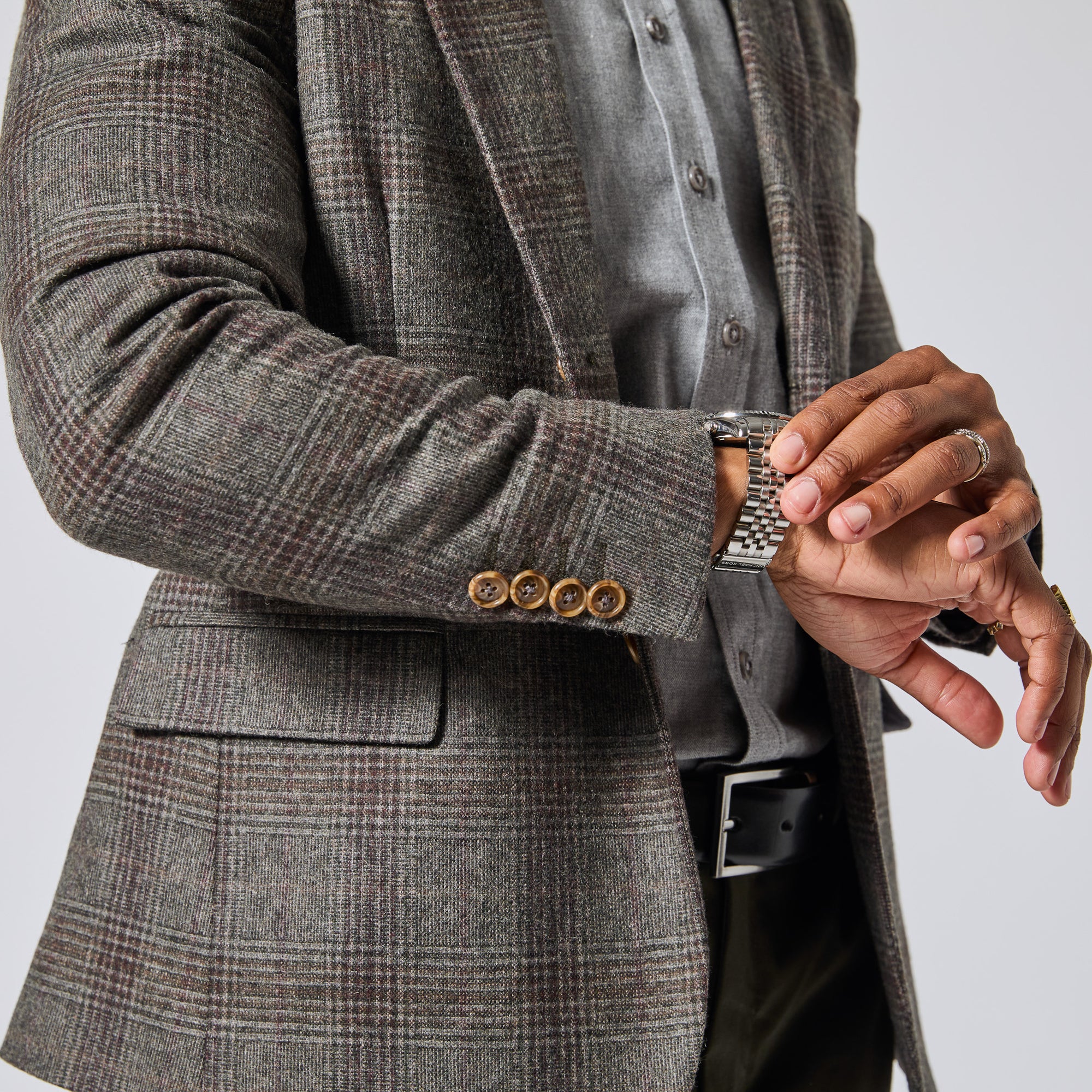 A close-up of a person wearing a gray and brown plaid blazer over a gray button-up shirt, with a silver metal link watch visible on the wrist. Color: Earthen Glen Plaid