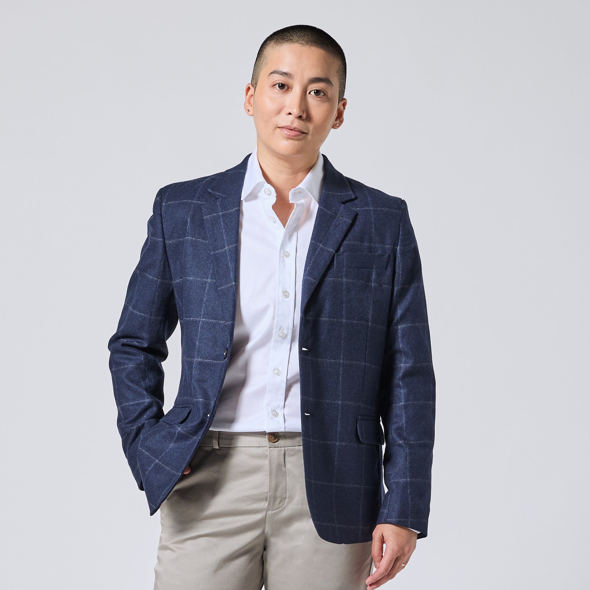Model wearing a navy blue windowpane plaid blazer over a white button-down shirt and beige trousers, standing against a light gray studio background. Color: Navy Regatta