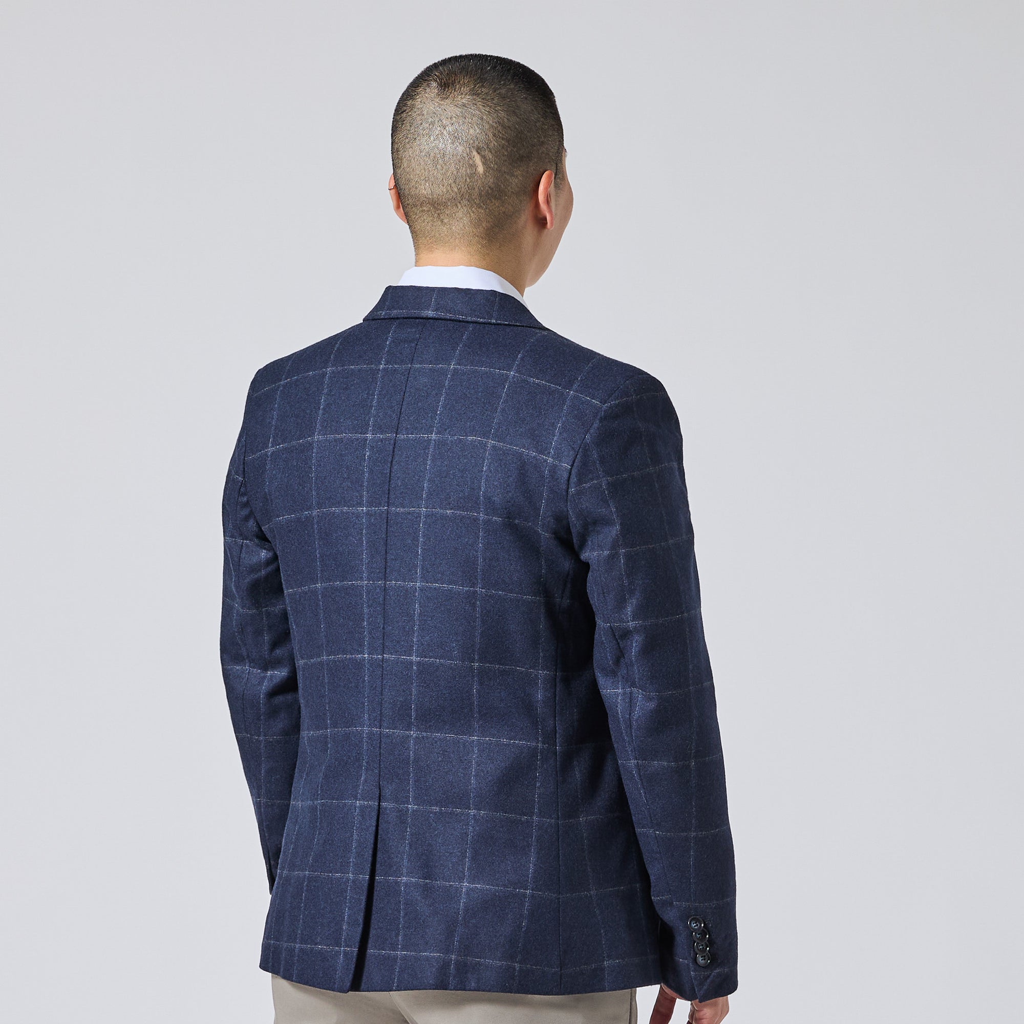 Model viewed from the back wearing a navy blue windowpane plaid blazer over a white collared shirt and beige trousers, set against a plain light gray studio background. Color: Navy Regatta