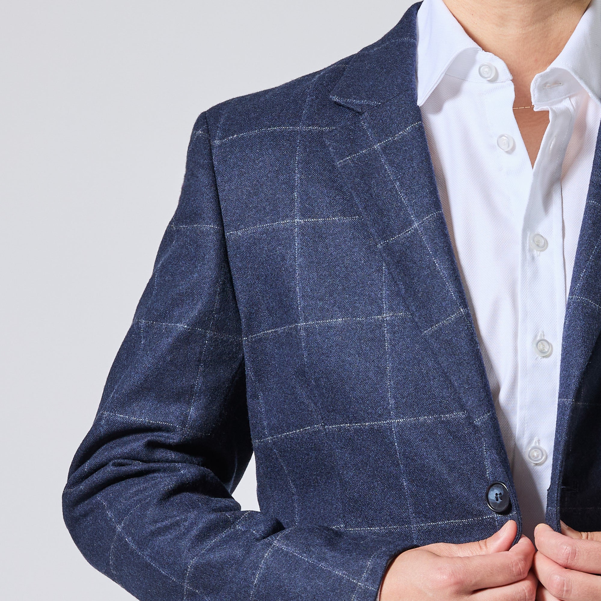 A close-up of a model wearing a navy blue windowpane plaid blazer over a white button-down shirt, with their hands adjusting the front button. Color: Navy Regatta