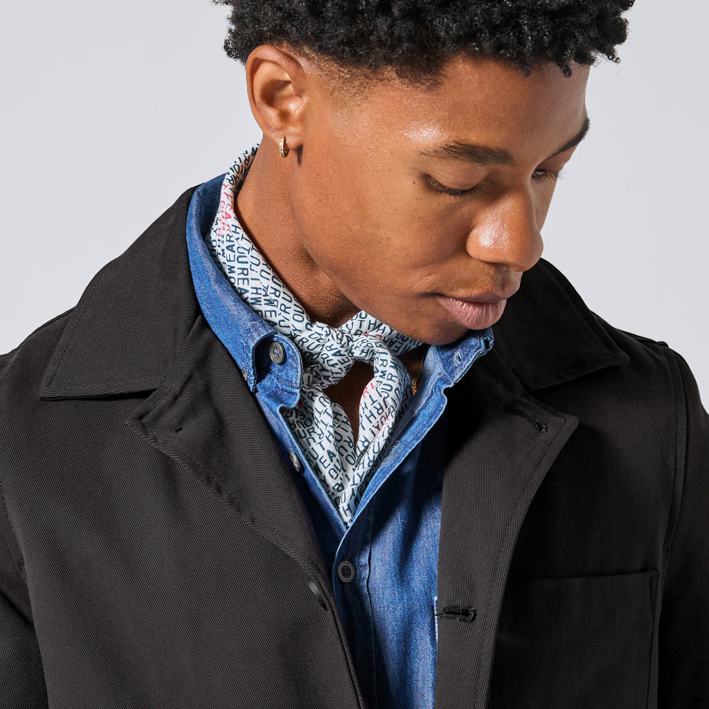 A close-up of a model wearing a black coat over a blue denim shirt and a patterned bandana tied around their neck looking down against a light gray background.