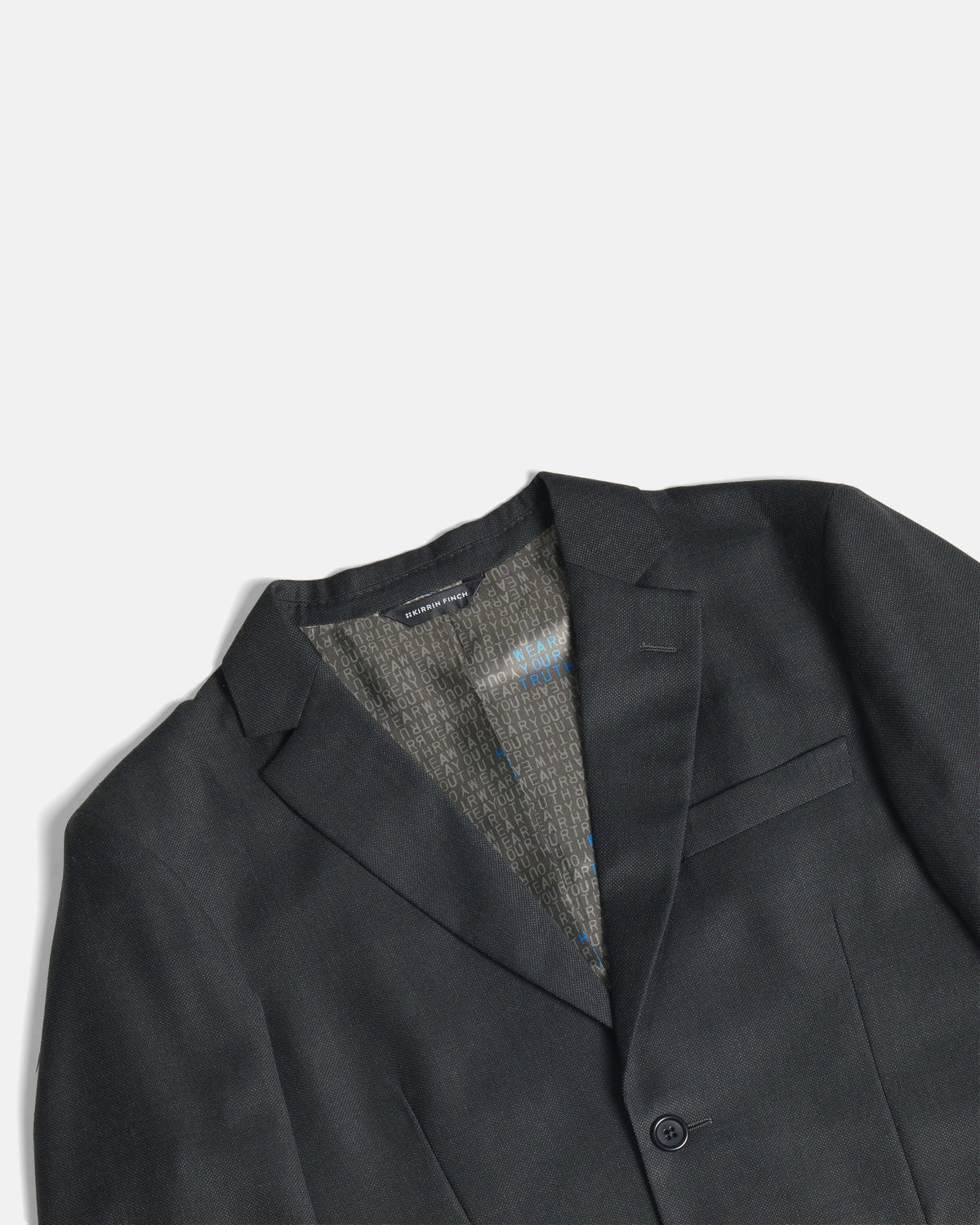 Close-up of a dark charcoal gray blazer with a textured gray and blue patterned lining visible inside the collar, set against a clean off-white background. Color: Nightfall Birdseye
