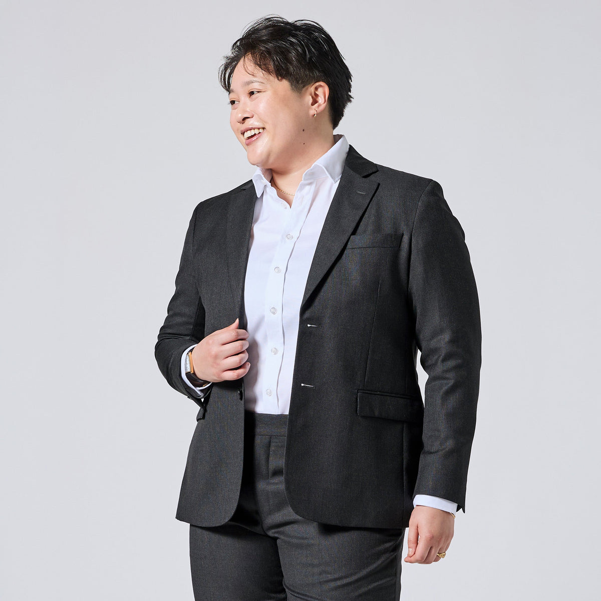 Person wearing a dark gray blazer over a white button-down shirt and dark gray trousers, smiling and looking to the side against a light gray studio background. Color: Nightfall Birdseye