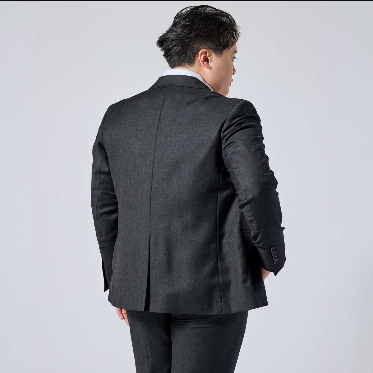 Person wearing a dark charcoal gray suit jacket, shown from the back and three-quarter side view against a light gray studio background. Color: Nightfall Birdseye