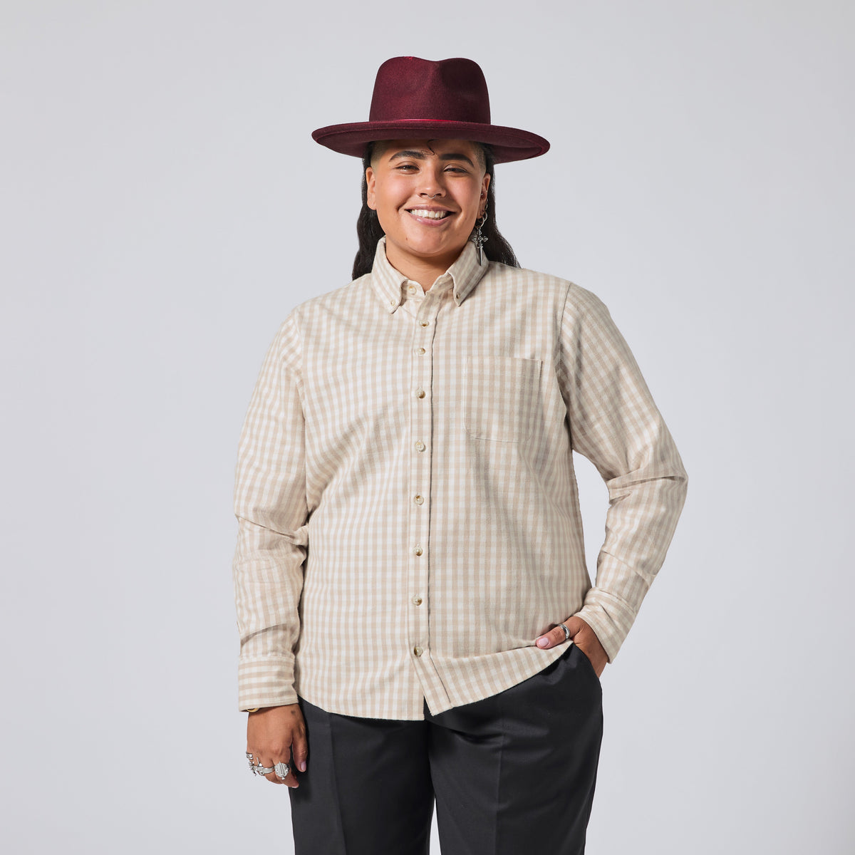 Model in a beige and white gingham long-sleeve button-down shirt, paired with dark gray pants and a burgundy wide-brim hat, against a solid light gray background. Color: Cream Check Flannel