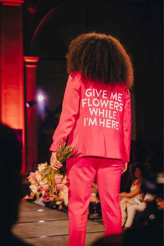 Runway model wearing a red suit with the words "Give me flowers while I'm here"