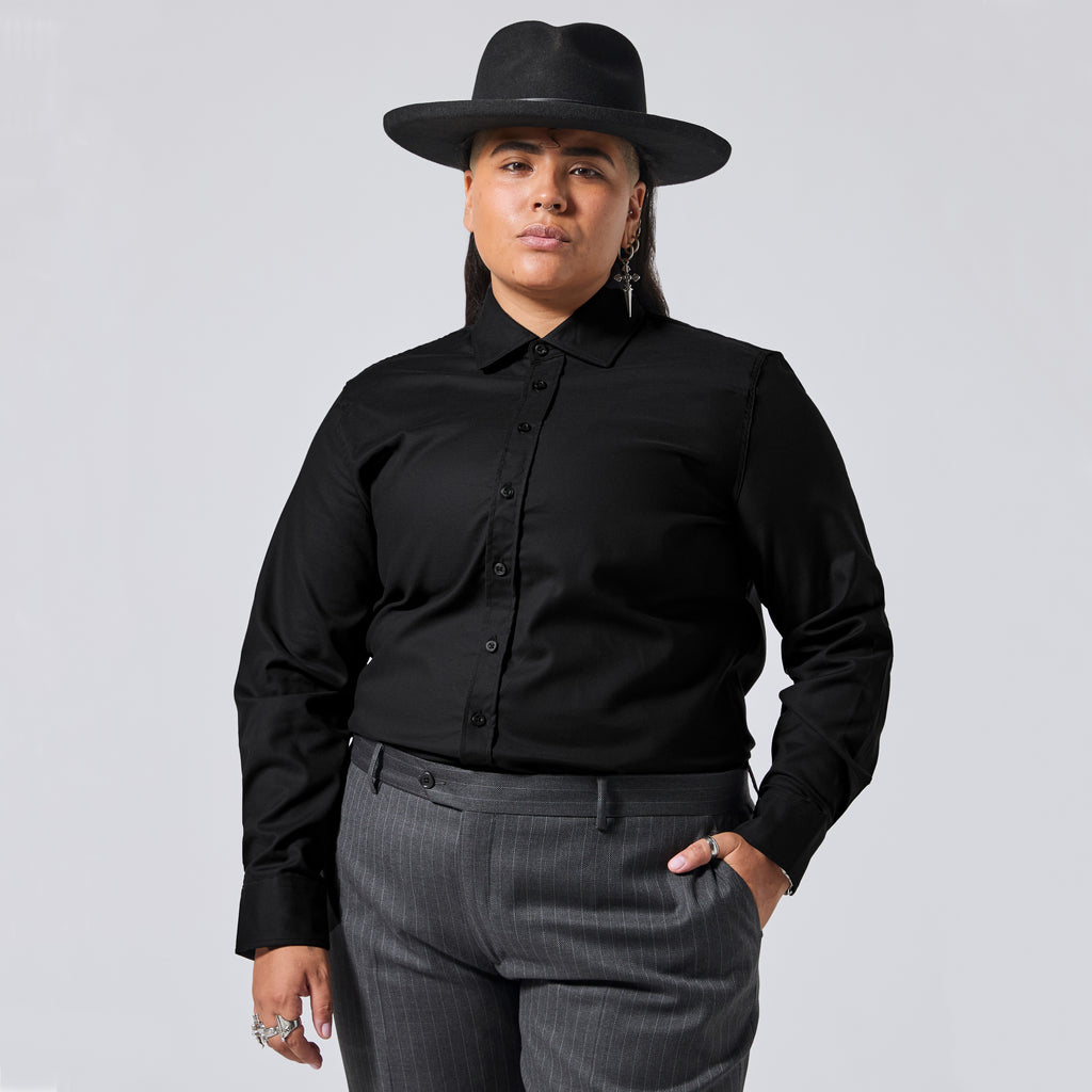A model wearing a black long-sleeve button-down shirt, a black wide-brim fedora hat, and dark gray pinstripe trousers, posed against a solid light gray background. Color: Black