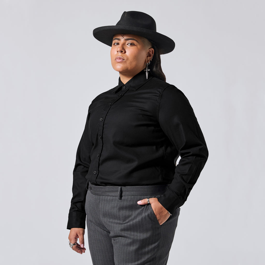 A model wearing a black long-sleeve button-up shirt, a black wide-brim hat, and gray pinstripe trousers, photographed against a solid light gray background. Color: Black