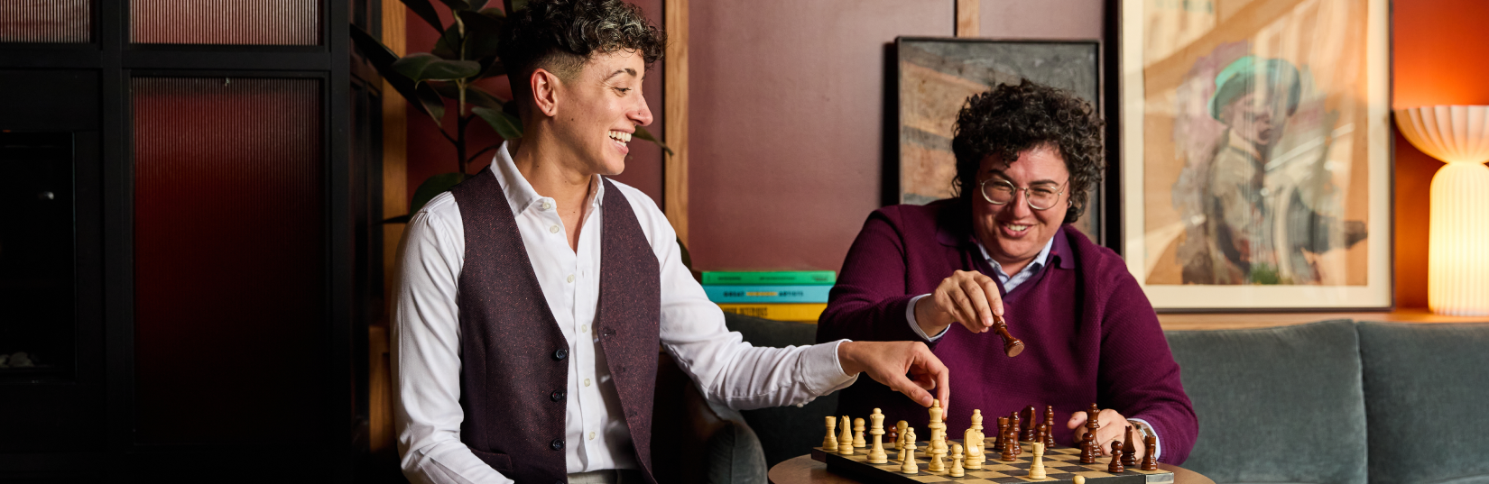 Two people playing chess in a cozy room with framed pictures on the wall.