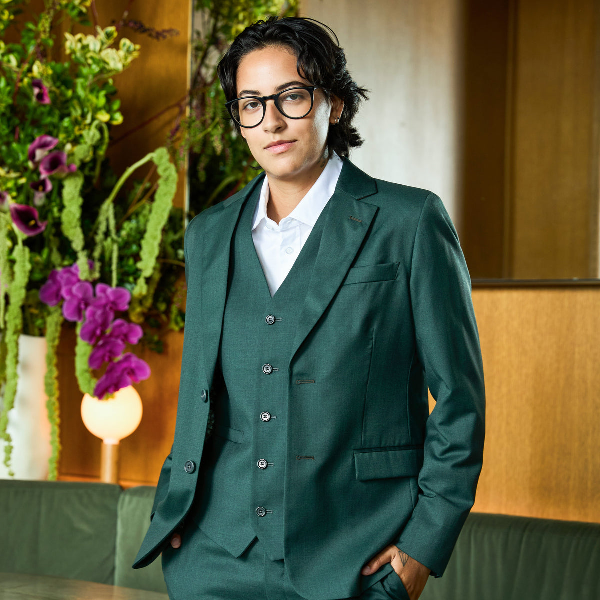 Model wearing a dark green three-piece suit with a white collared shirt, standing in a warm indoor setting with a floral arrangement in the background. Color: Emerald