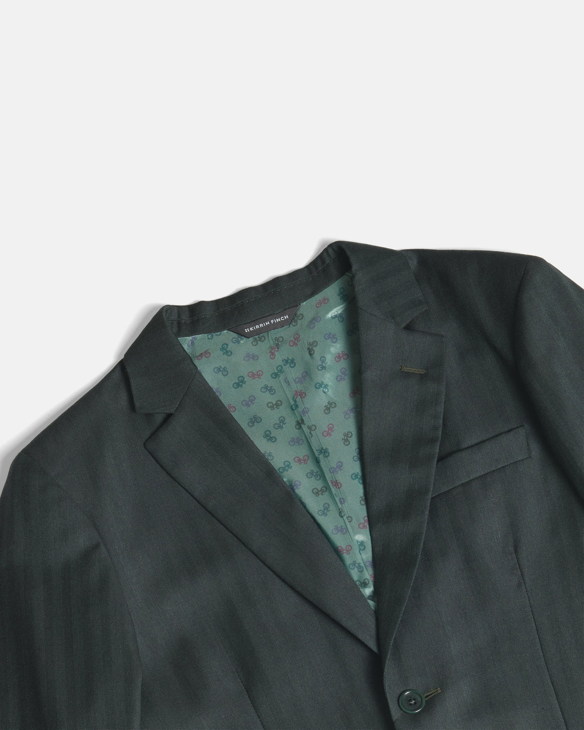A close-up of a dark green suit jacket, showing the textured fabric, notch lapels, and a patterned green inner lining. Color: Midnight Pine Herringbone