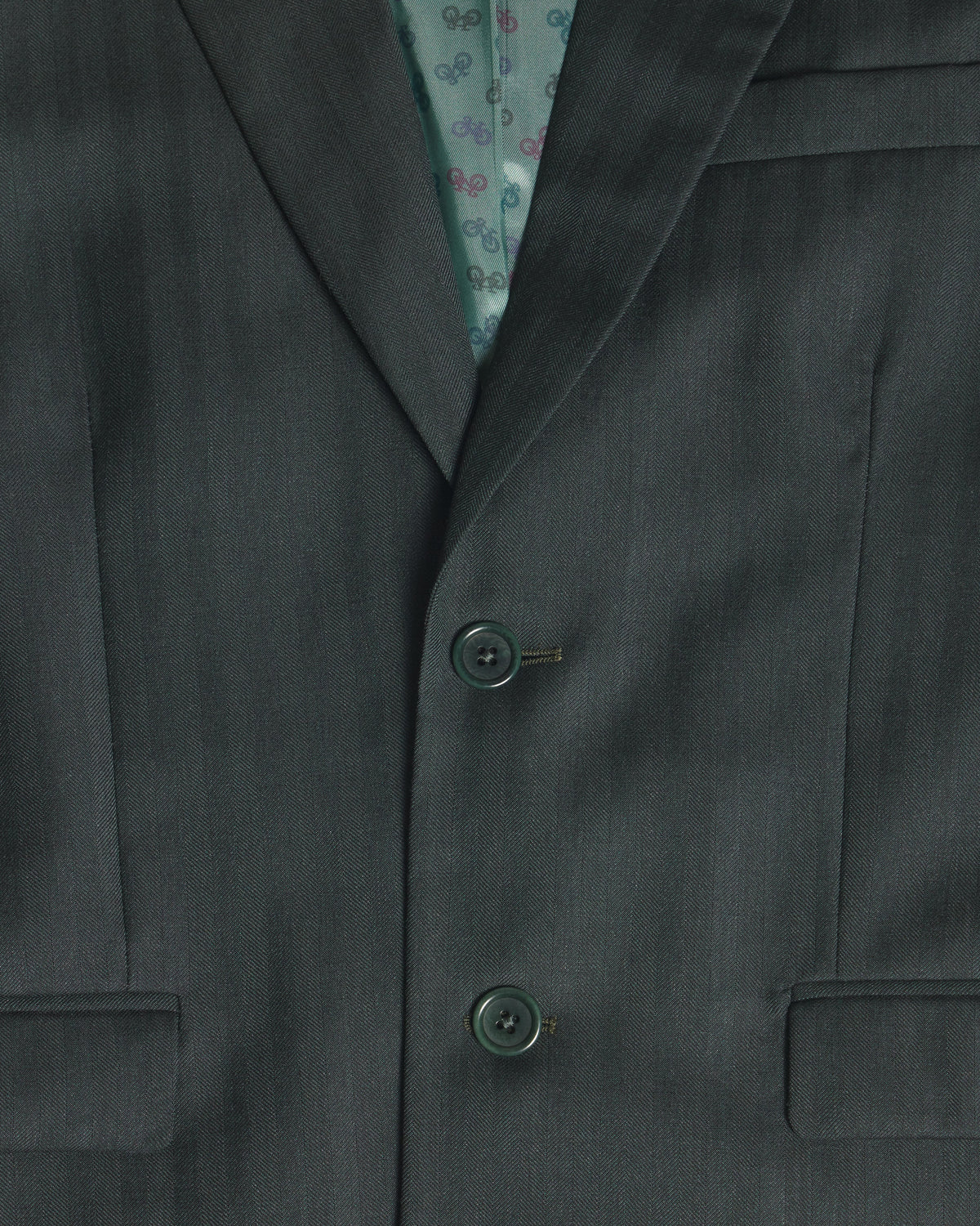 Close-up of a dark green suit jacket with two buttons, showing a patterned teal inner lining. Color: Midnight Pine Herringbone
