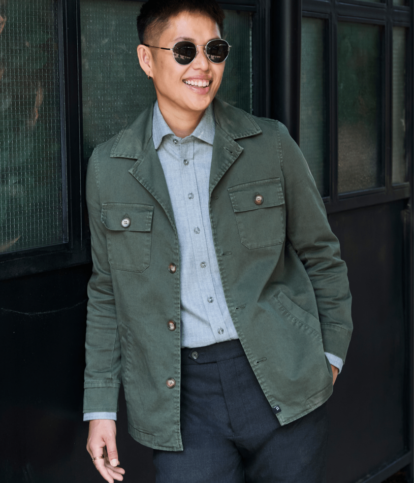 A smiling person wearing round sunglasses, a frankie button-up shirt, an olive green utility jacket, and dark trousers, standing in front of a dark building with window panes.
