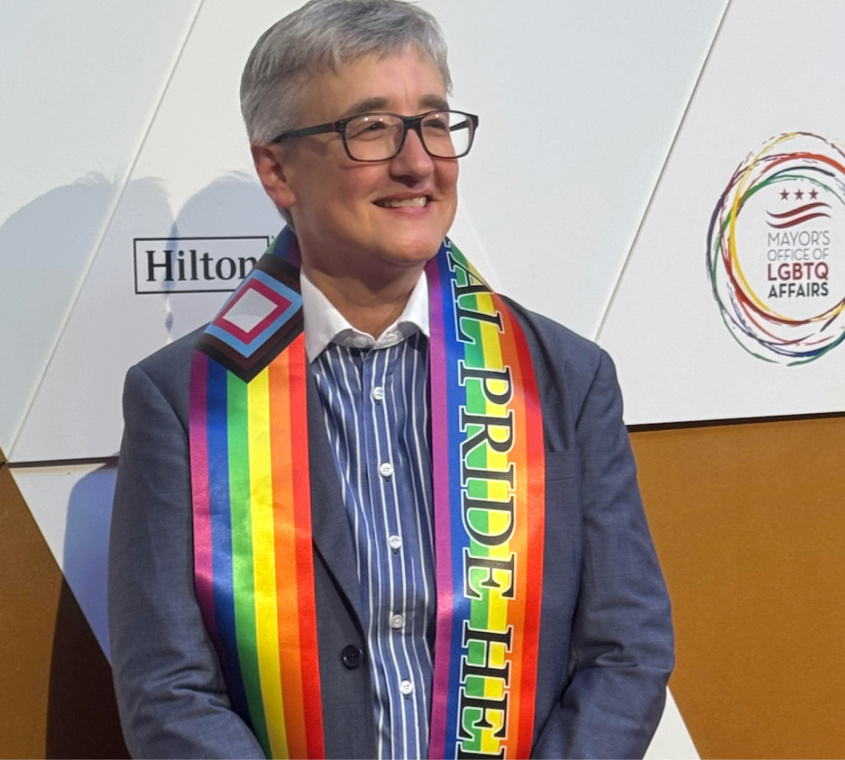 A smiling woman wearing glasses and a slate blue blazer over a striped banker shirt, with a rainbow pride sash that reads 'AL PRIDE HE' around her neck, standing in front of an event backdrop with Hilton and Mayor's Office of LGBTQ+ Affairs logos.