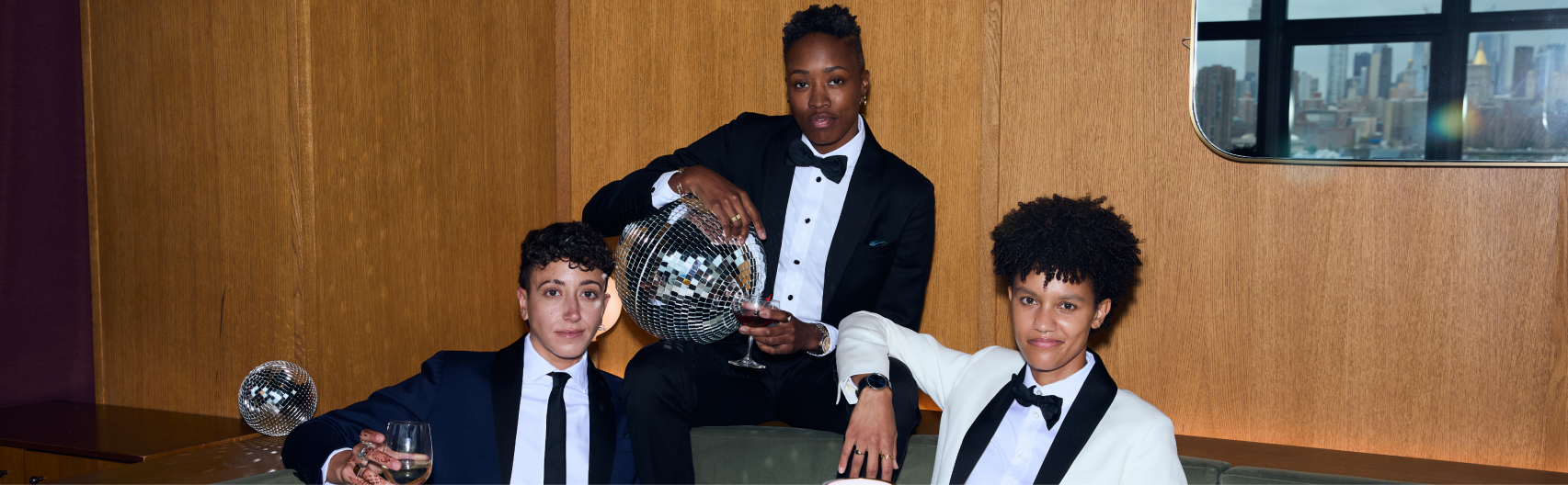 Three people sitting on a couch in formal wear holding drinks and a disco ball.
