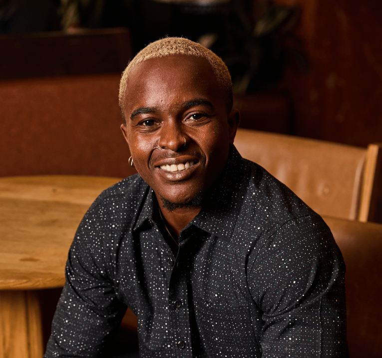 A smiling man with short blonde hair wearing a dark patterned button-up shirt, seated in a warm indoor setting with wooden furniture and brown leather seating visible in the background.