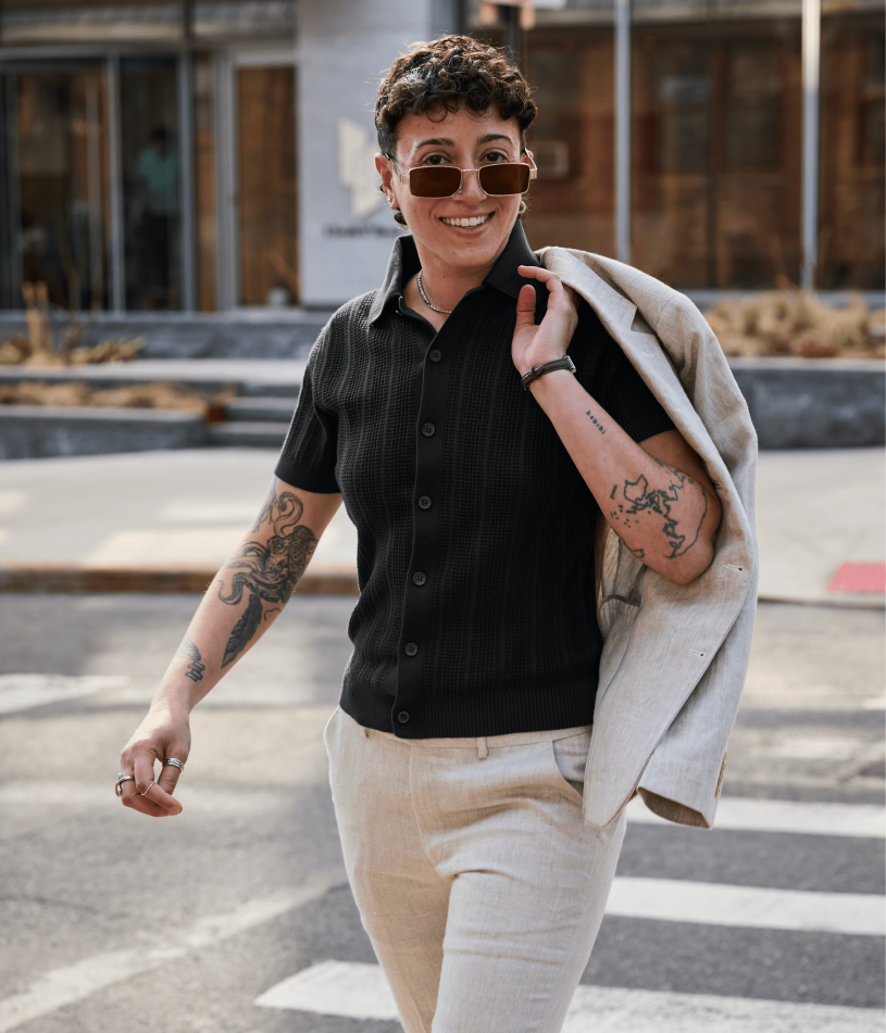 A woman walking outdoors wearing a black short-sleeve button-up knit cardigan, light beige linen pants, sunglasses, and carrying a light beige blazer over her shoulder.