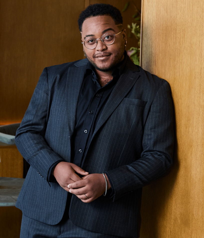 A man wearing a dark pinstripe suit jacket over a black button-down shirt, leaning against a light wood-paneled wall.