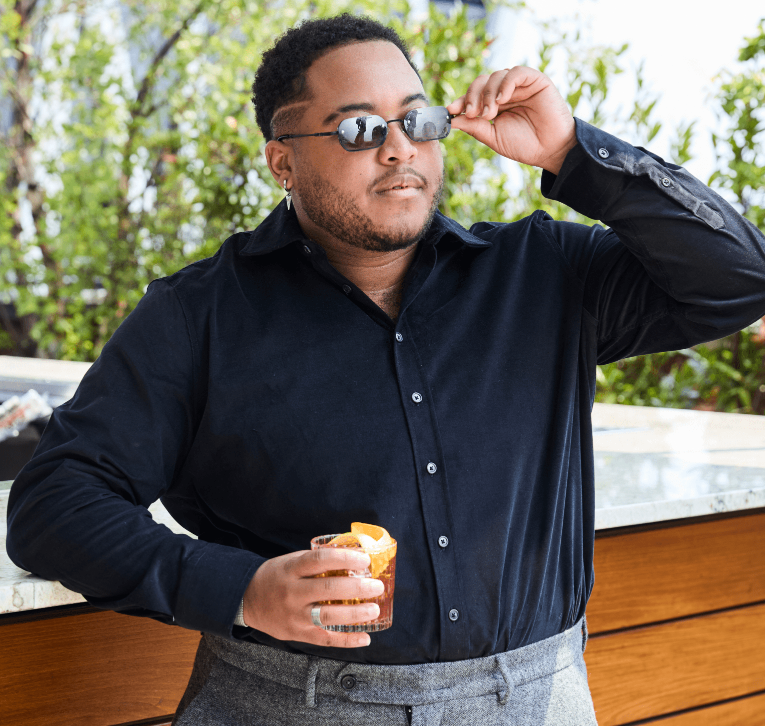 A man wearing a black long-sleeve button-up shirt and gray trousers, holding a glass of orange-colored drink outdoors while adjusting his sunglasses.