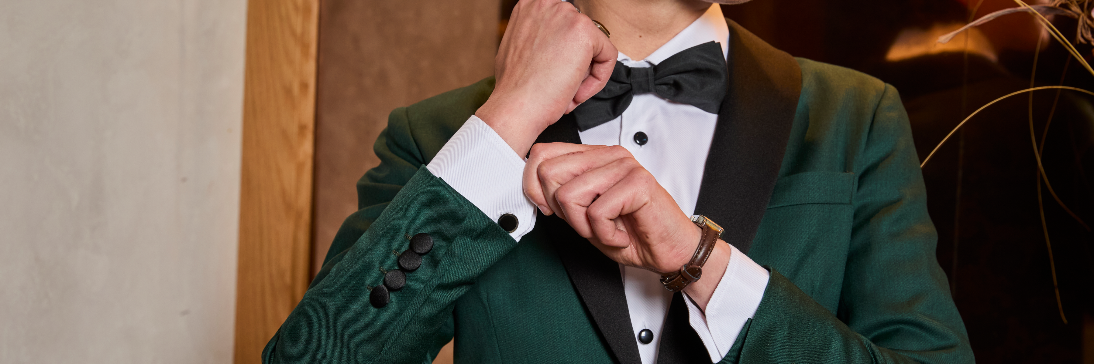 Person adjusting a cuff in a formal emerald tuxedo with a neutral background.