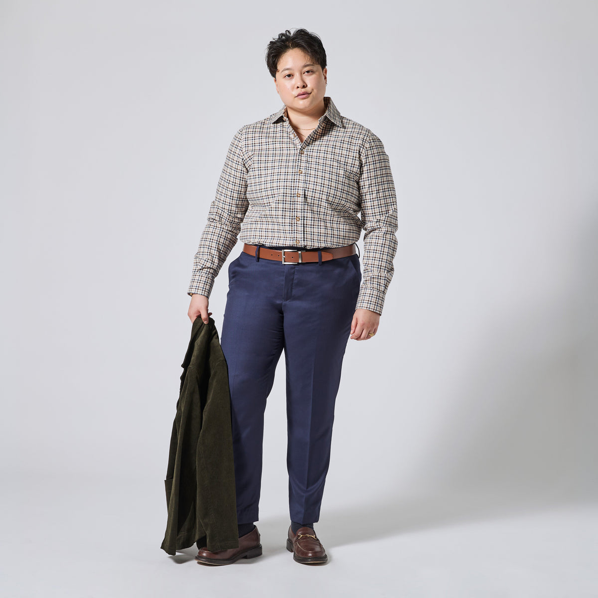 Person modeling a long-sleeve button-up plaid shirt in beige, brown, and gray, paired with navy blue trousers, a brown leather belt, and brown shoes. They are holding an olive green jacket against a light gray studio background. Color: Herritage Plaid