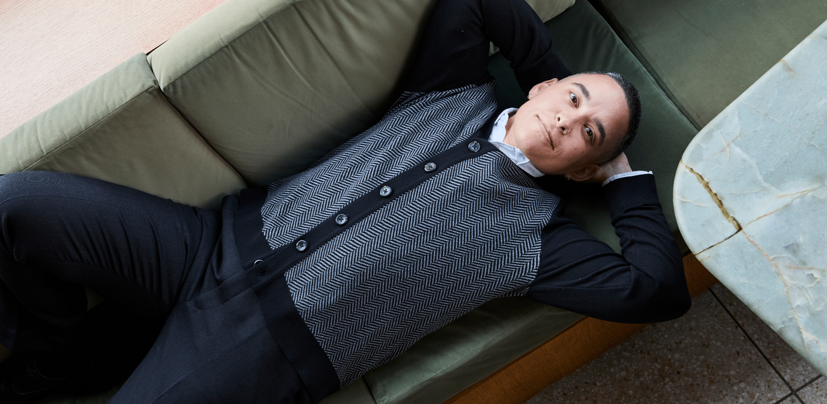A person wearing a black-and-gray herringbone cardigan with black trim, reclining on a green sofa.