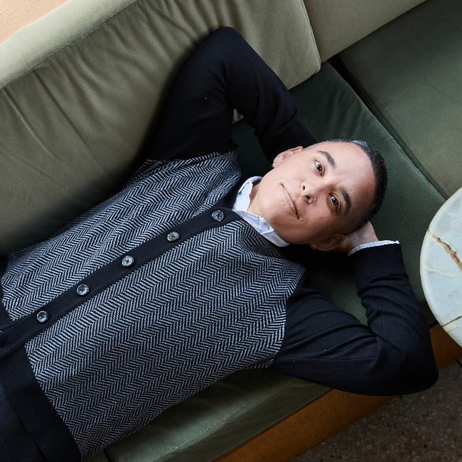 A person wearing a black-and-gray herringbone cardigan with black trim, reclining on a green sofa.