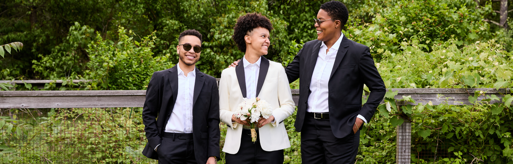 Three people in formal attire standing outdoors with greenery in the background