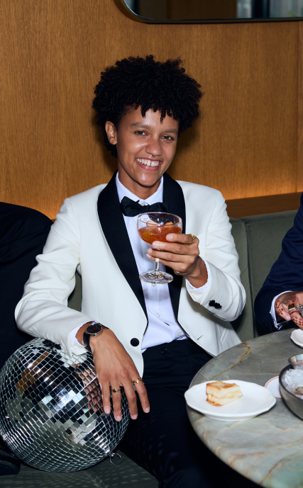 Person in ivory Kirrin tuxedo sitting down with a drink in hand leaning on a disco ball 