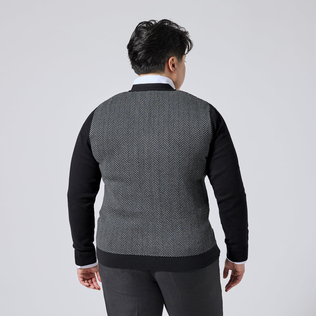 Model viewed from the back wearing a long-sleeve knit sweater with a black and white herringbone pattern on the body and solid black sleeves, collar, and hem, paired with dark trousers against a light gray studio background. Color: Inkstone Herringbone