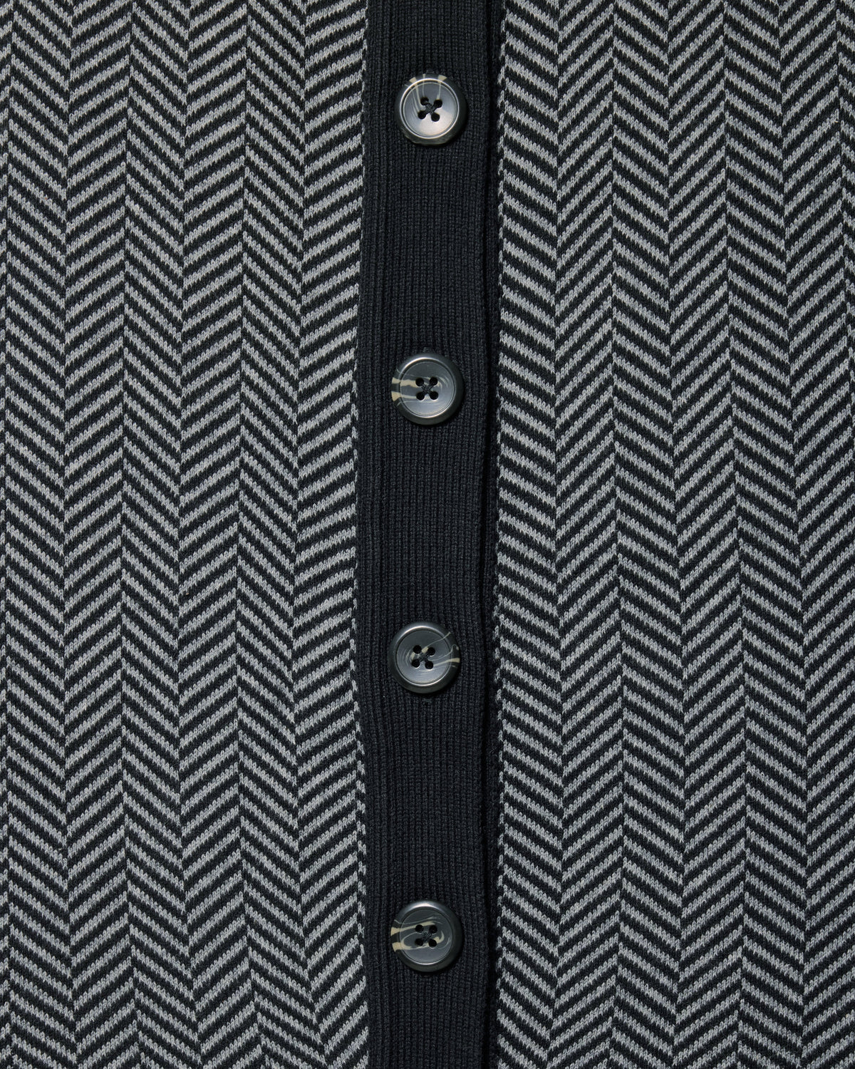 Close-up of a herringbone patterned knit garment in dark gray and black, featuring a solid black center placket with five dark buttons. Color: Inkstone Herringbone