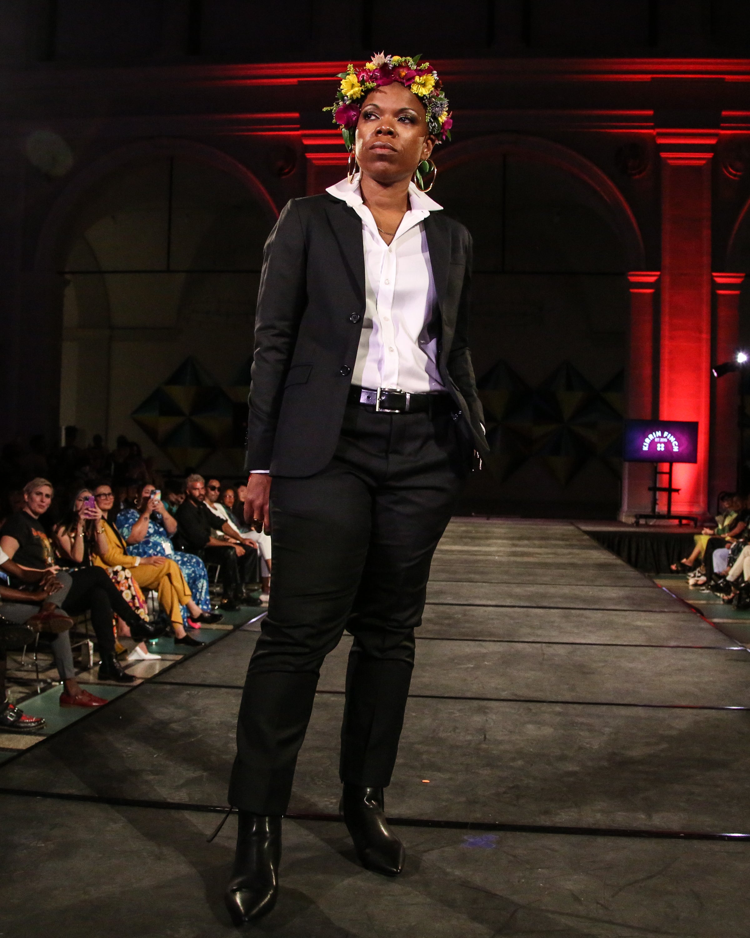 Model in a black suit and flower crown walking the runway.