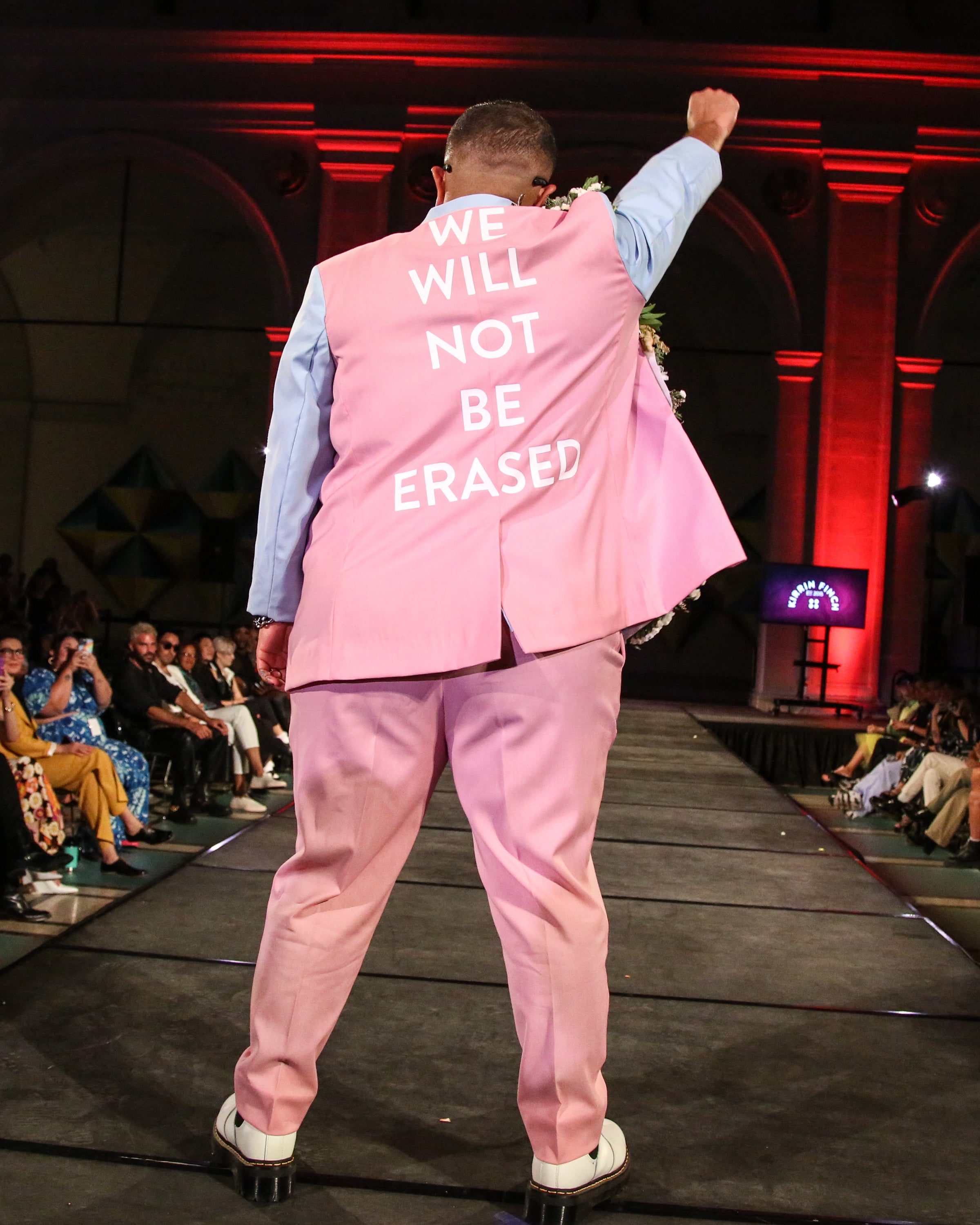 Runway moderl wearing a pink and blue blazer with the words " We will not be erased" on the back of it.