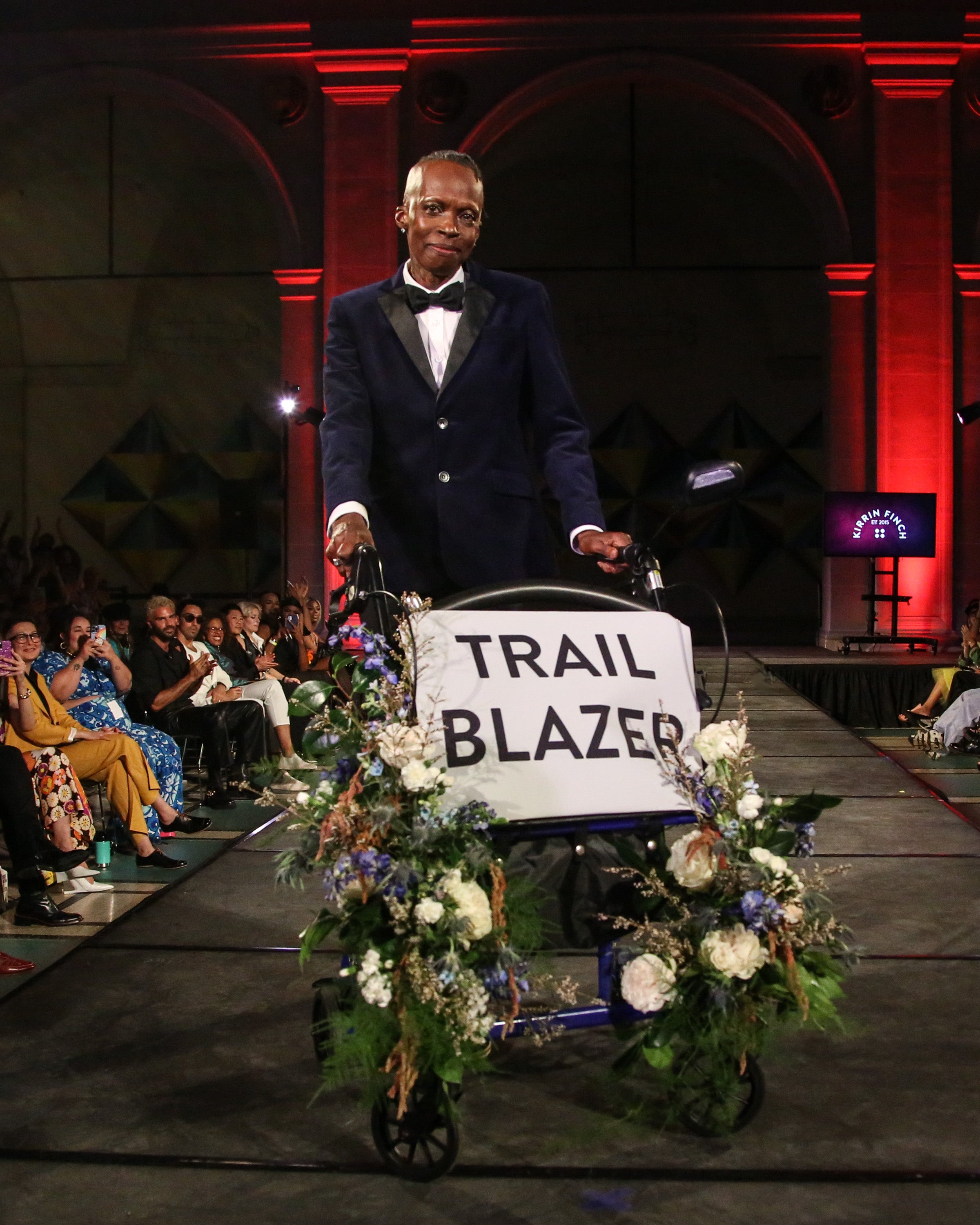 Model in dark blue tuxedo walking runway with a rollator adorned with flowers.