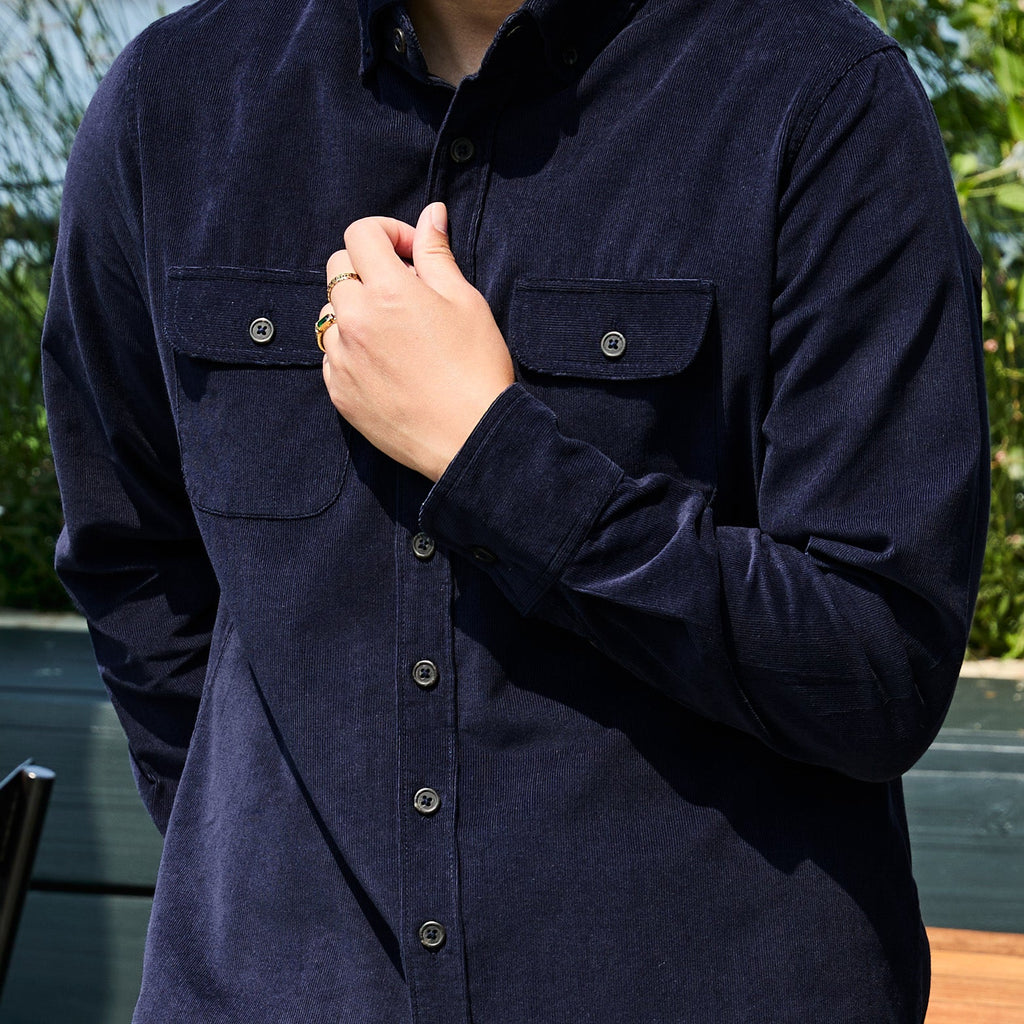 A close-up of a person wearing a dark navy corduroy long-sleeve button-up shirt with two chest pockets and metal buttons, set against an outdoor background with green foliage. Color: Maritime Blue