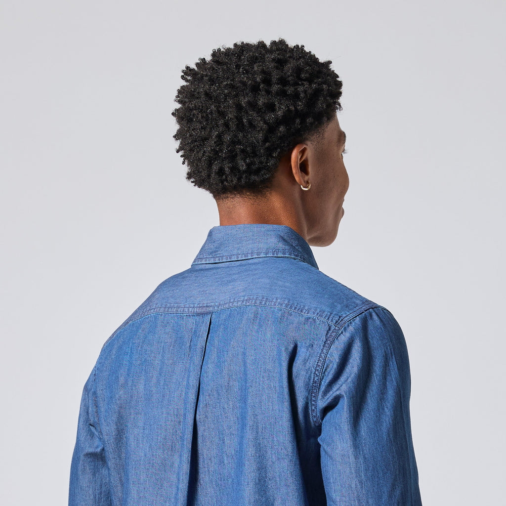 A person shown from the back wearing a blue denim button-up shirt against a light gray studio background. Color: Medium Wash Denim