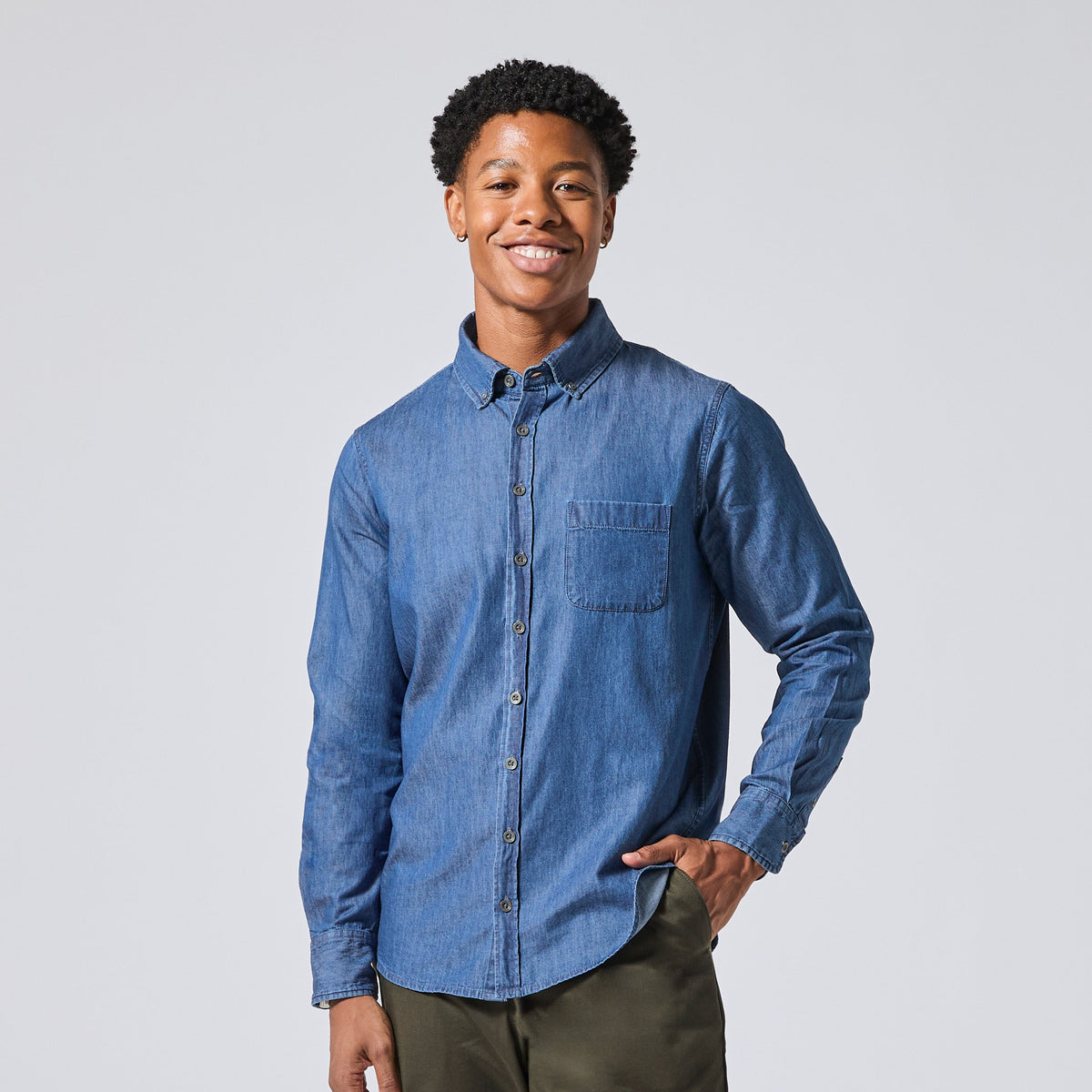 A smiling model wearing a medium blue long-sleeve button-down shirt with a chest pocket, paired with olive green pants, standing against a light gray studio background. Color: Medium Wash Denim