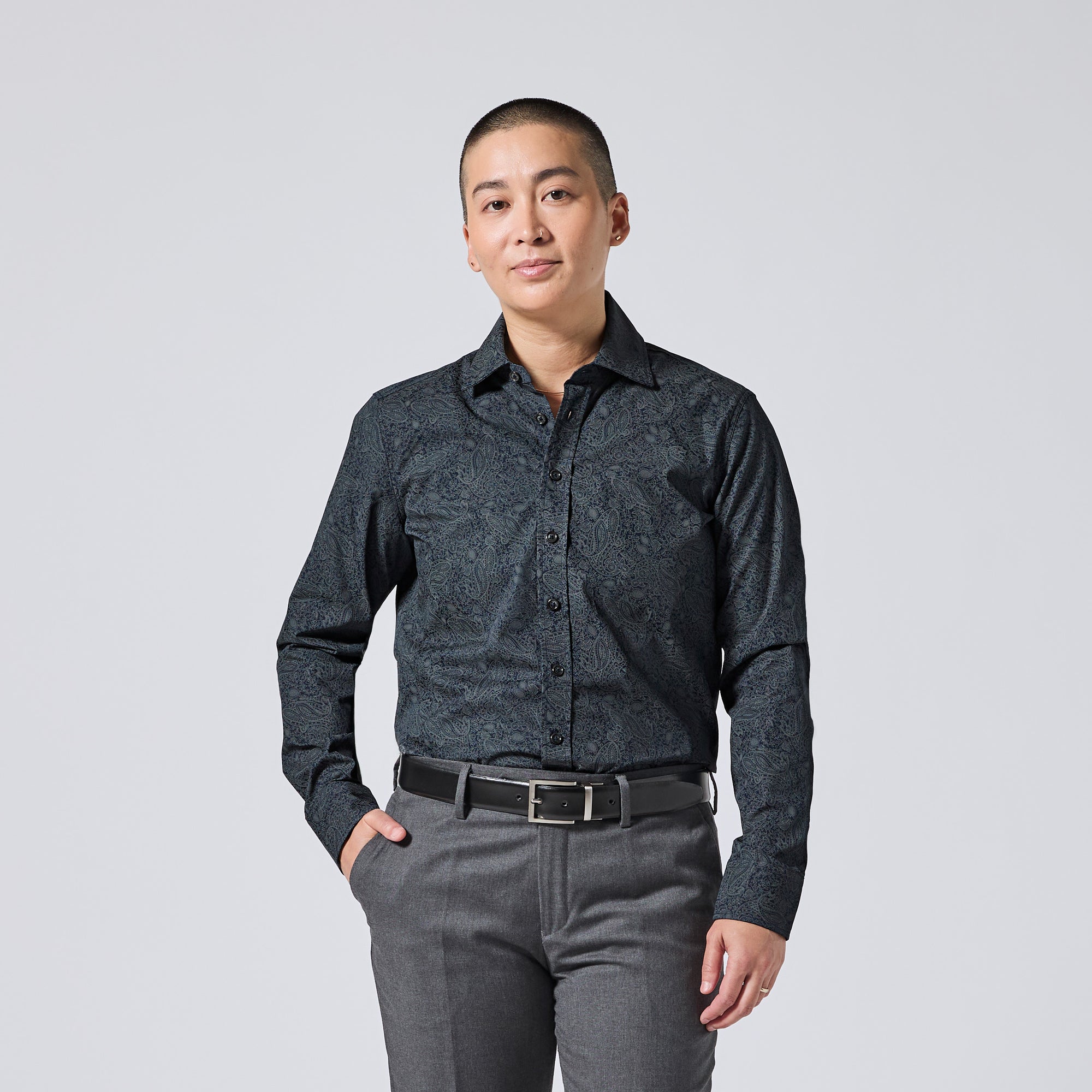 Model wearing a dark charcoal long-sleeve button-down shirt with a subtle tonal paisley pattern, paired with gray trousers and a black belt, standing against a light gray studio background. Color: Midnight Paisley