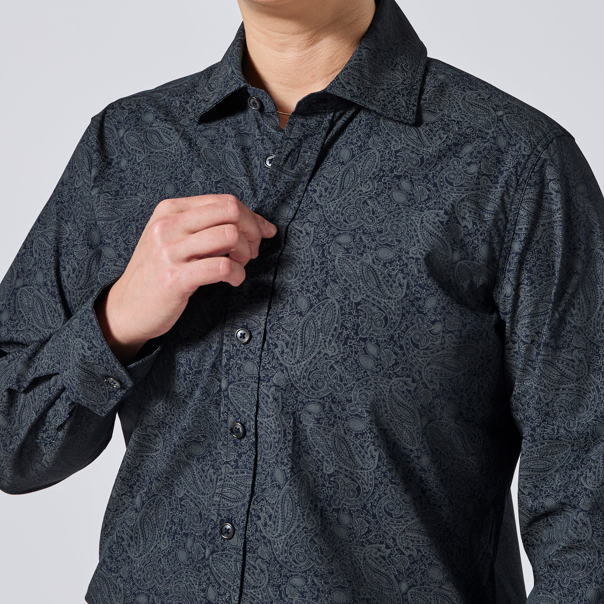 A close-up of a model wearing a dark charcoal gray long-sleeve button-down shirt with a subtle paisley pattern. They are adjusting the collar with their right hand, and the background is a clean, neutral light gray studio backdrop. Color: Midnight Paisley