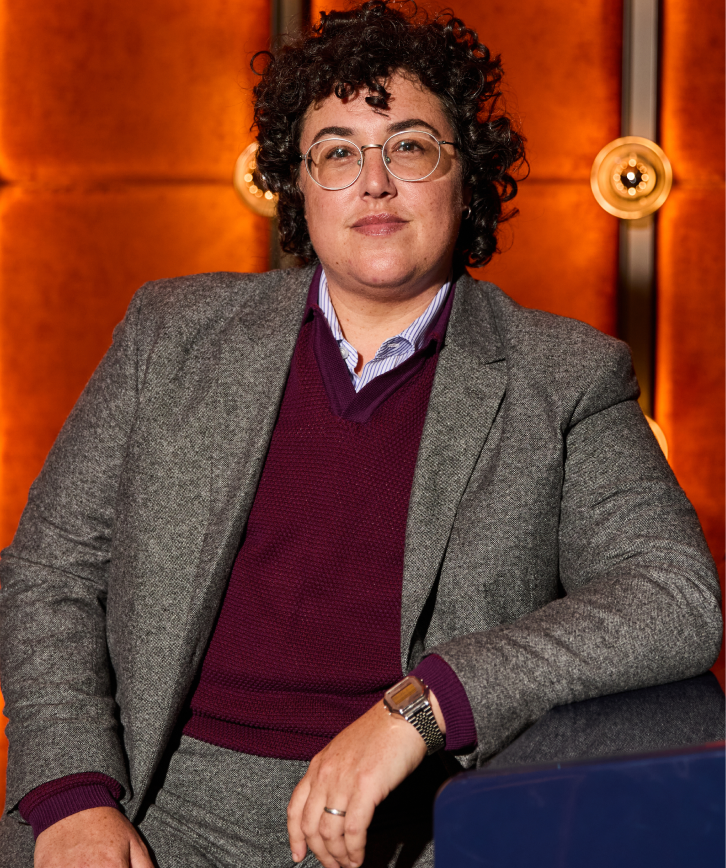 Person wearing a gray blazer and maroon sweater against an orange wall.