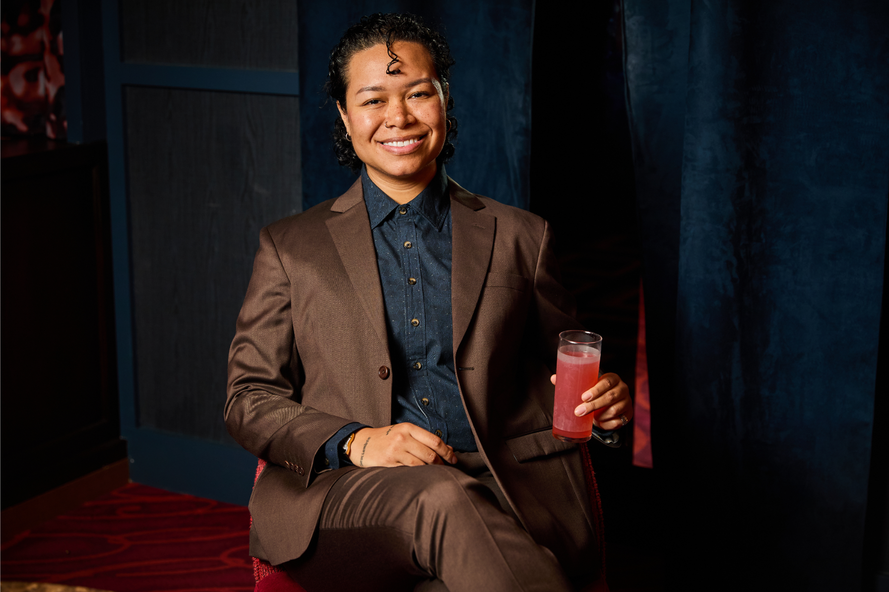 Person in a brown suit holding a pink drink against a dark background