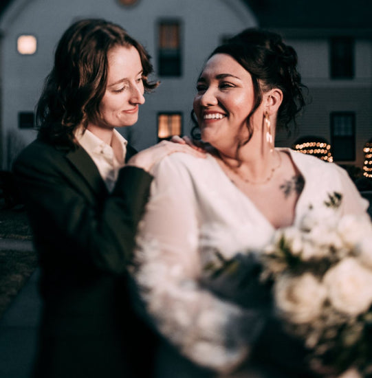 Newlywed couple embracing with one holding a bouquet, standing in front of a building with lights.