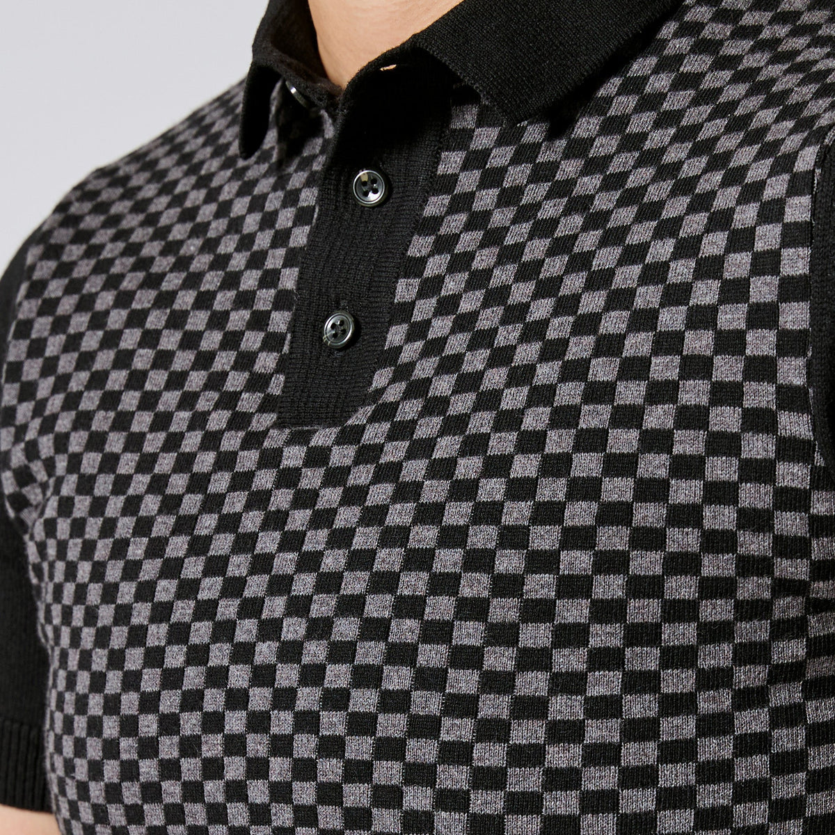 Close-up of a black and gray checkered knit polo shirt with a black collar and two dark buttons. Color: Shadow Checker